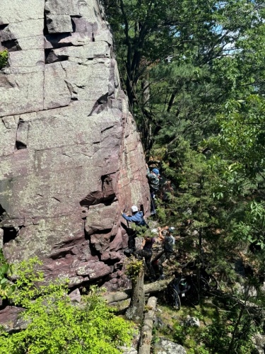 Several climbers, wearing helmets and harnesses, partway up a steep purple quartzite rock face while others assist near the base.