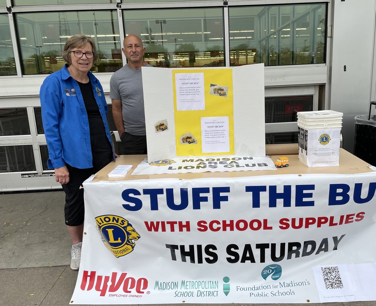 A woman in a blue jacket and a man in a gray shirt stand behind a table with a 'Stuff the Bus' banner